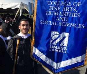 UMass Lowell Alum, Nicholas Gates stands next to the FAHSS banner at Commencement.