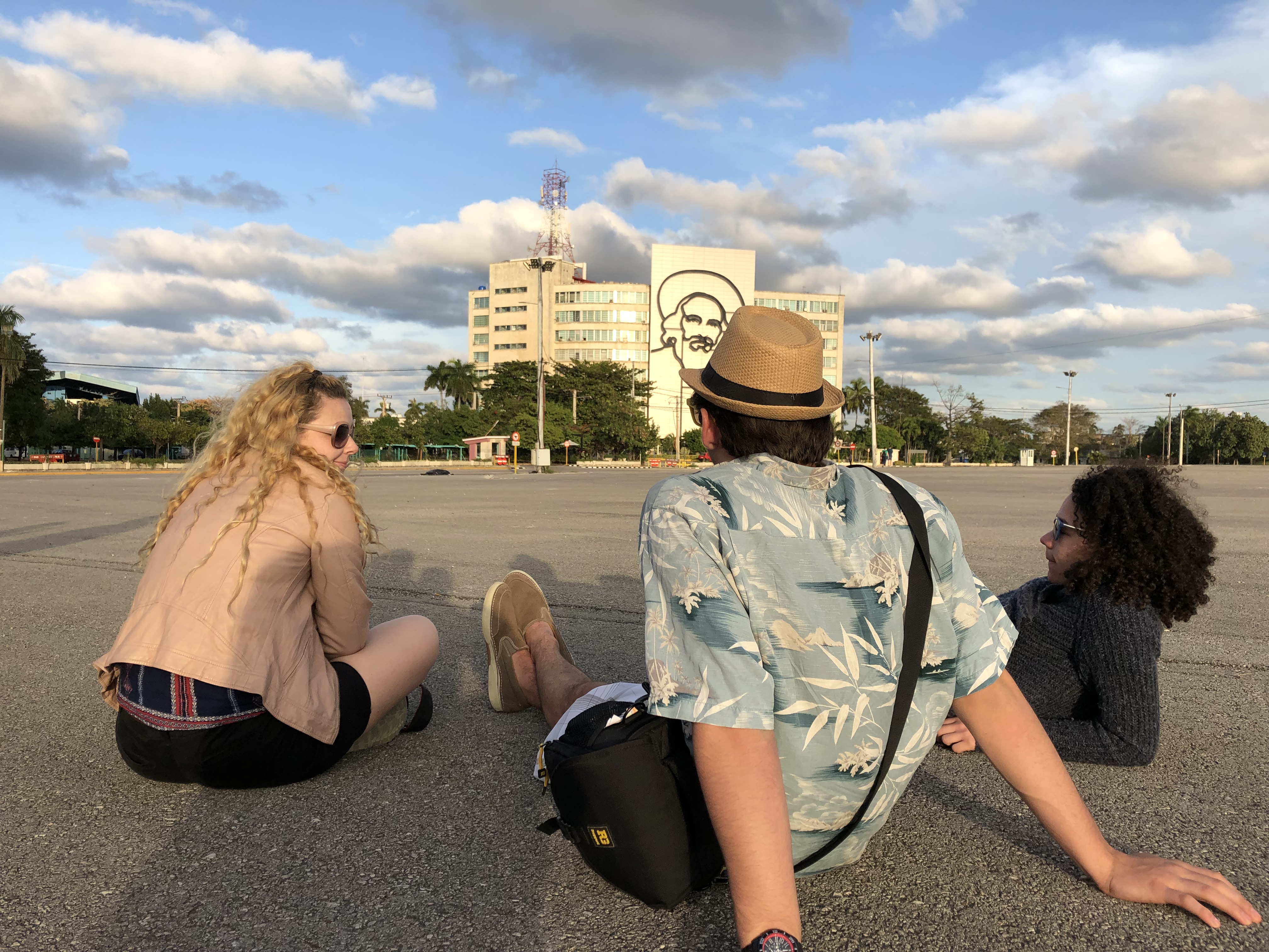 Three honors students sit in Revolution Square in Havana, looking out at the portrait of Cienfuegos on a building.