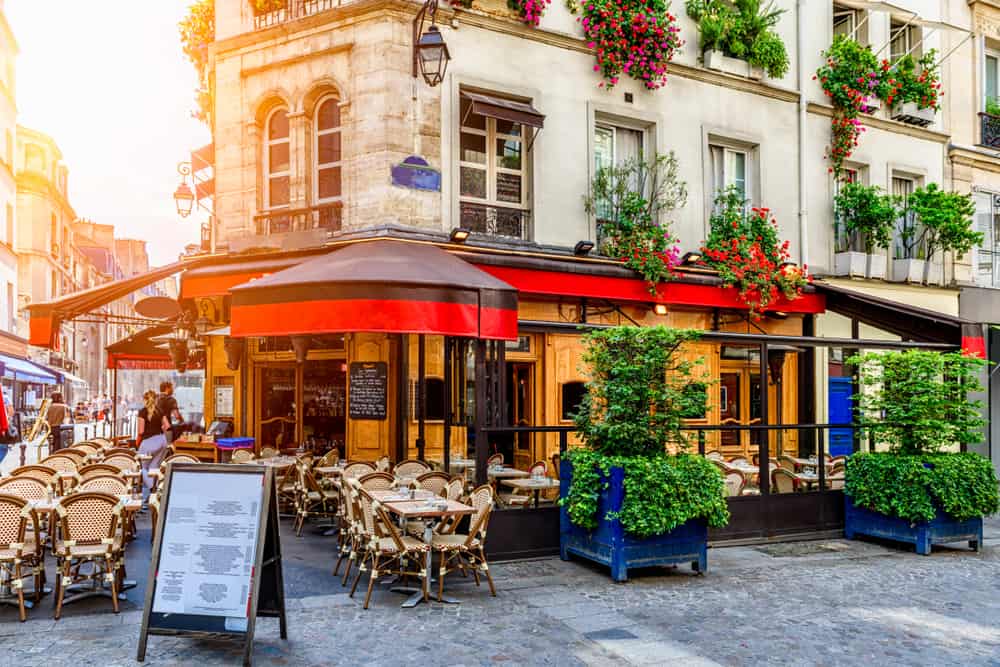 A beautiful Parisian cafe with red awning and outdoor seating, while, in the left corner, a dusky golden light shines.