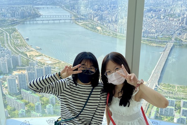 Trang Vy Bui and another woman pose for a, picture holding up peace signs with a city scape behind them.