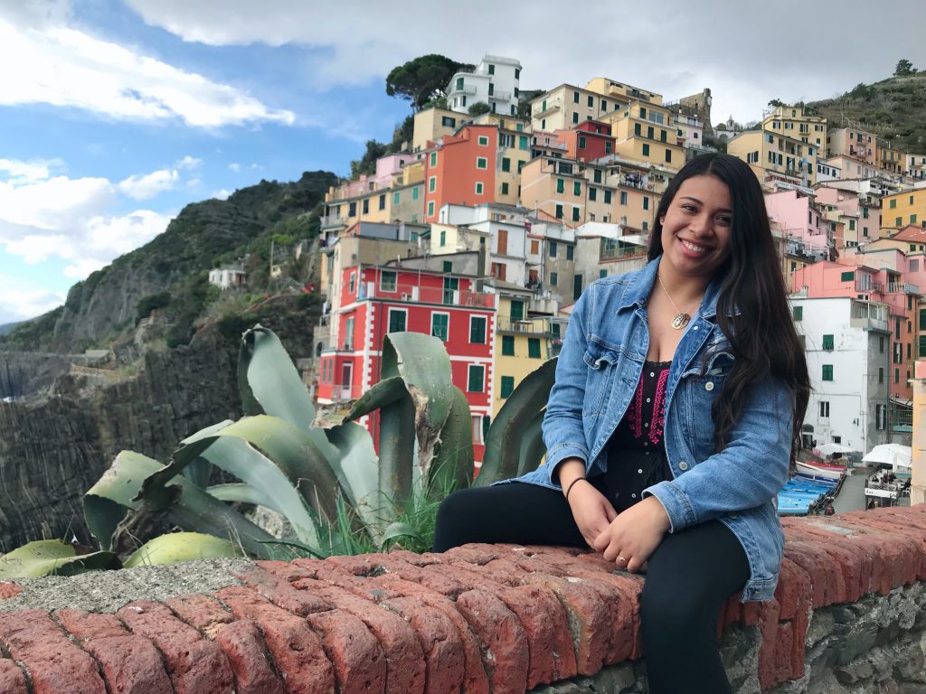 Kelsey Gonzalez-sitting on a brick wall in front of multi-colored homes built into the side of a mountain